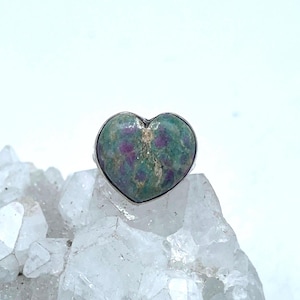 May include: A heart-shaped ring with a green and purple gemstone set in a silver band. The stone has a mottled appearance. The ring is displayed on a cluster of clear quartz crystals. The ring is a piece of jewellery.