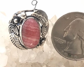 May include: A sterling silver ring with an oval, pink rhodochrosite stone. The ring features floral and leaf designs around the stone. A US quarter is next to the ring for size comparison.
