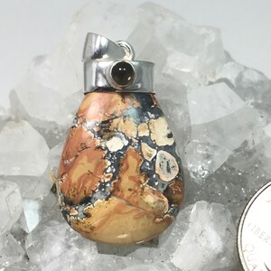 May include: A teardrop-shaped pendant with a silver bail. The stone features a mix of tan, brown, black, and white patterns. A small, round, dark-colored stone is set in the silver top. The pendant is displayed on a bed of clear crystals.