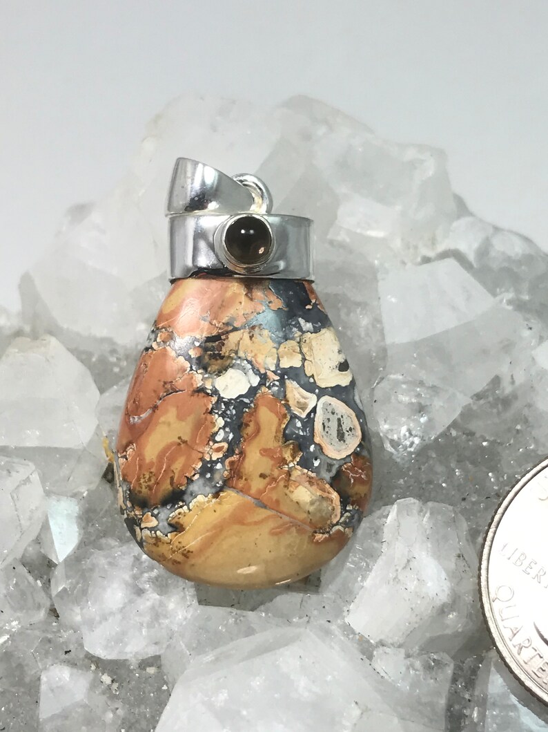 May include: A teardrop-shaped pendant with a silver bail. The stone features a mix of tan, brown, black, and white patterns. A small, round, dark-colored stone is set in the silver top. The pendant is displayed on a bed of clear crystals.