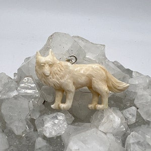 Könnte beinhalten: Ein weißer, geschnitzter Wolf-Anhänger, mit einer silbernen Öse zum Anbringen an einer Halskette oder einem Armband. Der Wolf steht auf einem Bett aus klaren Quarzkristallen.