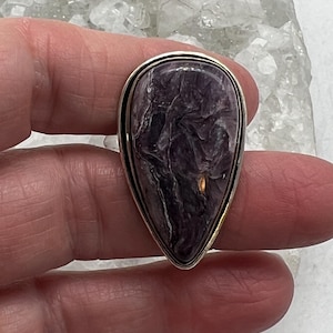 May include: A teardrop-shaped ring featuring a polished, marbled purple stone set in a silver-tone metal frame with a gold-tone inner rim. The stone's pattern includes swirls of dark purple and white.