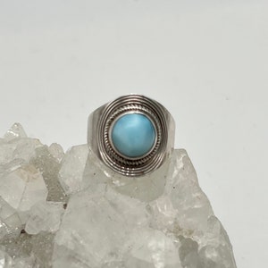 May include: A silver ring featuring a round, light blue gemstone. The gemstone is set within a circular design of concentric lines and a rope-like border. The ring is displayed on a rough, crystalline surface.