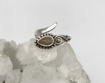 Op de afbeelding: Een zilveren ring met een heldere kwarts steen gezet in een gedetailleerd ontwerp. De ring ligt op een bed van witte kristallen.