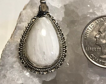 May include: A teardrop-shaped pendant with a white stone set in a silver-colored, ornate frame. The pendant is next to a U.S. quarter for size comparison. The frame has a detailed, beaded design around the stone.