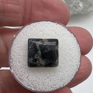 May include: A square, polished gemstone with a black and white marbled pattern. The stone is set in a clear plastic container with a white, textured interior. The gemstone is held in a person's hand.