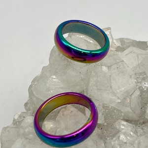 May include: Two iridescent rings with a rainbow color scheme, including shades of purple, blue, green, and yellow. The rings are displayed on a white, crystalline surface. The rings have a smooth, polished finish.