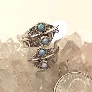 Può includere: Anello color argento con design a forma di piuma, con tre pietre di opale blu chiaro. L'anello è presentato su uno sfondo di cristalli trasparenti, con una moneta per riferimento.