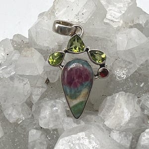 May include: A silver pendant with a teardrop-shaped, multi-colored stone, featuring red, blue, and green hues. The pendant is adorned with three green peridot stones and a small red gemstone. The pendant is suspended from a silver bail.