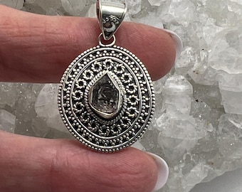 May include: A silver pendant with an intricate, circular design. The pendant features a central, irregularly shaped, clear gemstone. The silver setting has a detailed, openwork pattern around the gemstone and a decorative border. The bail is also silver.