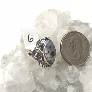 Pu&ograve; includere: Un anello d'argento con una pietra ovale bianca e grigia incastonata in una fascia d'argento con un design a spirale. L'anello &egrave; su un letto di cristalli bianchi. Il numero "6" &egrave; scritto su un'etichetta bianca.