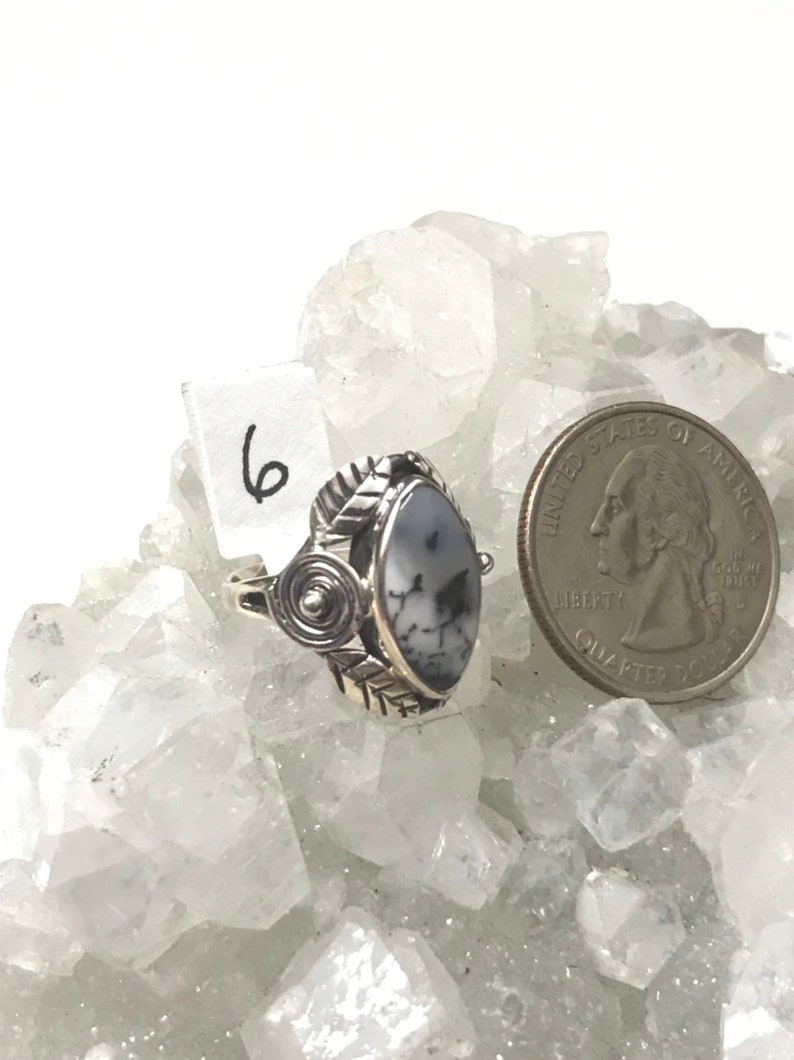 Pu&ograve; includere: Un anello d'argento con una pietra ovale bianca e grigia incastonata in una fascia d'argento con un design a spirale. L'anello &egrave; su un letto di cristalli bianchi. Il numero "6" &egrave; scritto su un'etichetta bianca.