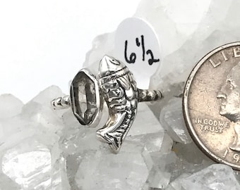 Op de afbeelding: Zilveren ring met een visontwerp en een heldere, rechthoekige steen. De ringmaat is 6 1/2. De ring wordt getoond op een wit, kristallijn oppervlak, met een Amerikaanse kwart dollar munt ter vergelijking.