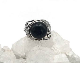 May include: A silver ring featuring a large, round, black gemstone. The ring has an intricate, decorative design around the stone, with a leaf pattern. The ring is displayed on a white, crystalline surface.