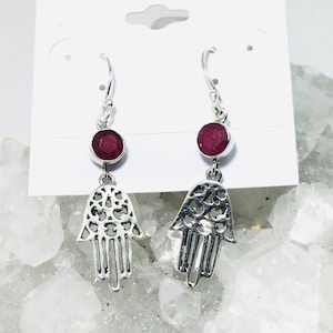 May include: Sterling silver earrings with a hamsa hand design and a red gemstone. The earrings are on a white card with the text "Sterling Silver".