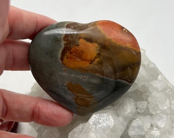 Puede incluir: Una piedra pulida en forma de corazón con tonos grises, marrones y anaranjados. La piedra se sostiene en una mano y descansa sobre un grupo de cristales de cuarzo transparente. El objeto es decorativo.
