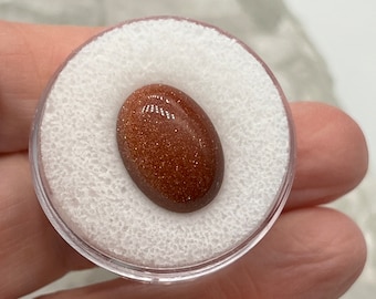 May include: A polished, oval-shaped gemstone with a sparkling, golden-brown appearance. The stone is set in a clear, round plastic container with a white foam insert. The gemstone is held in a person's hand.