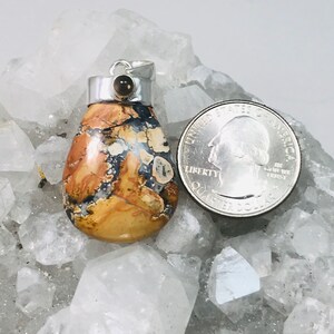 May include: A teardrop-shaped pendant with a silver bail and a small, round, brown stone. The pendant features a mix of tan, brown, and blue colors. A quarter is next to the pendant for size comparison. The pendant is on a white, crystal-like surface.