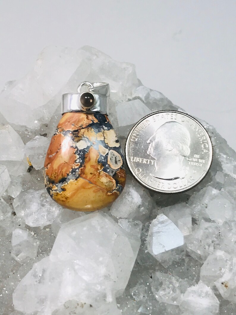 May include: A teardrop-shaped pendant with a silver bail and a small, round, brown stone. The pendant features a mix of tan, brown, and blue colors. A quarter is next to the pendant for size comparison. The pendant is on a white, crystal-like surface.