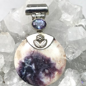 May include: A round, white and purple stone pendant with a silver bail and a small purple gemstone. The pendant is set on a bed of white crystals.