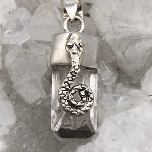 May include: A silver pendant featuring a clear quartz crystal with a silver snake design. The pendant is on a white background.