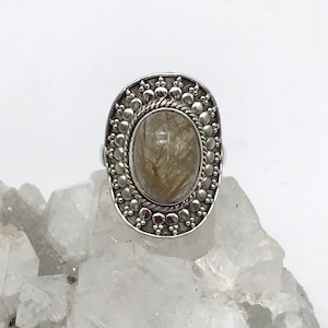 May include: Silver ring featuring an oval clear quartz stone with gold inclusions. The ring has a detailed silver band with a beaded design.