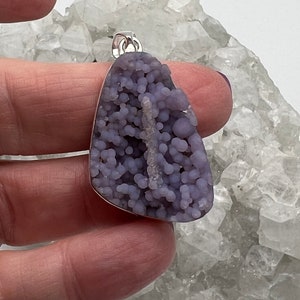 May include: A pendant featuring a unique, grape-like cluster of lavender-colored stone. The stone is set in a silver-colored metal frame with a small loop for a chain. The pendant is held against a backdrop of a white, crystalline surface.