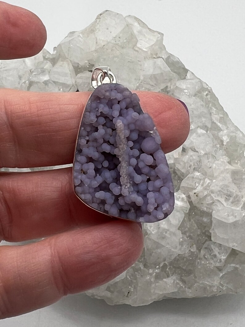 May include: A pendant featuring a unique, grape-like cluster of lavender-colored stone. The stone is set in a silver-colored metal frame with a small loop for a chain. The pendant is held against a backdrop of a white, crystalline surface.