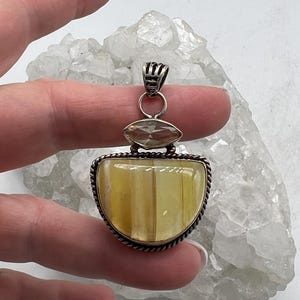 May include: A silver pendant with a large, semi-circular yellow gemstone, accented by a smaller, oval-shaped stone above. The pendant has a decorative silver border and a loop for a chain. The background is a white, crystalline surface.