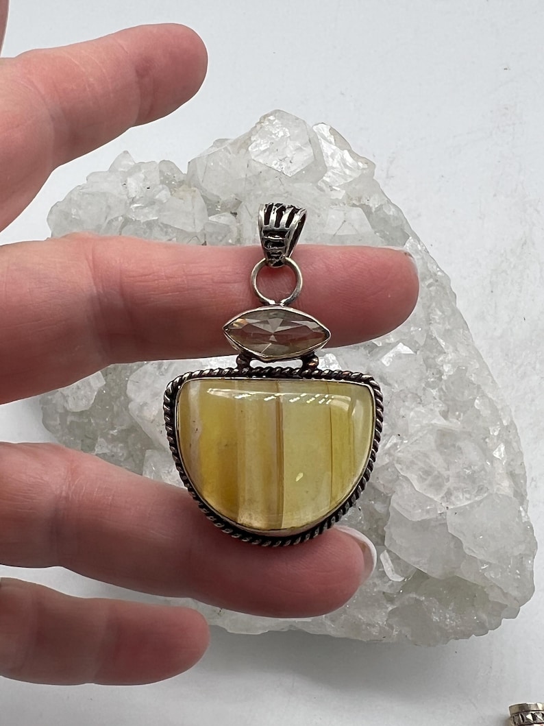 May include: A silver pendant with a large, semi-circular yellow gemstone, accented by a smaller, oval-shaped stone above. The pendant has a decorative silver border and a loop for a chain. The background is a white, crystalline surface.