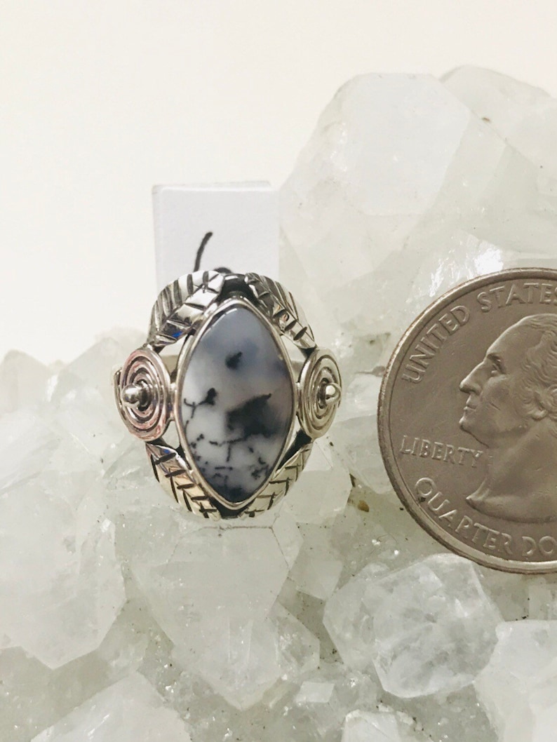 Pu&ograve; includere: Un anello d'argento con una pietra di agata dendritica bianca e nera incastonata in un design a spirale. L'anello &egrave; circondato da una fascia d'argento con un motivo a foglie.