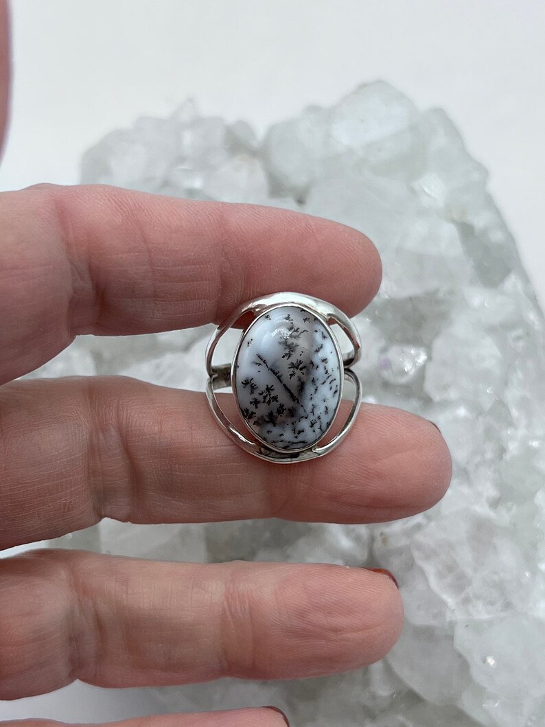 May include: A silver ring with a white and black dendritic agate stone. The stone is oval shaped and set in a silver band.