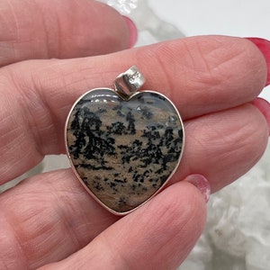 May include: Heart-shaped pendant with a silver bezel. The pendant features a natural stone with a pattern of black and tan inclusions. The bail is silver and stamped with "925".