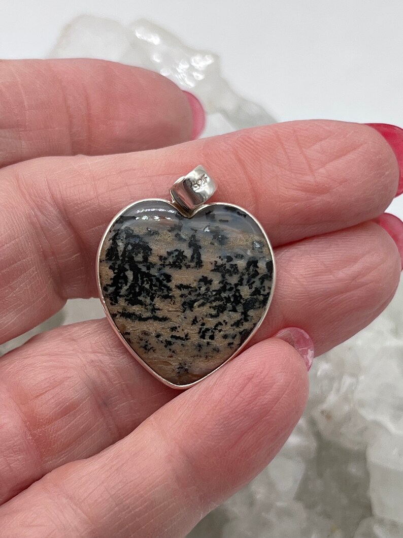 May include: Heart-shaped pendant with a silver bezel. The pendant features a natural stone with a pattern of black and tan inclusions. The bail is silver and stamped with "925".