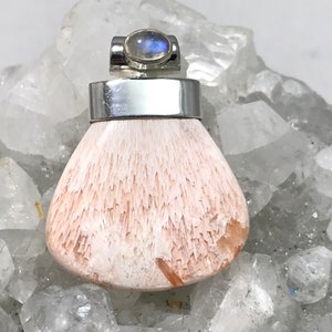 Op de afbeelding: Een zilveren hanger met een witte en roze edelsteen. De edelsteen is gezet in een zilveren bezel en heeft een moonstone cabochon bovenop. De hanger ligt op een bed van witte kristallen.