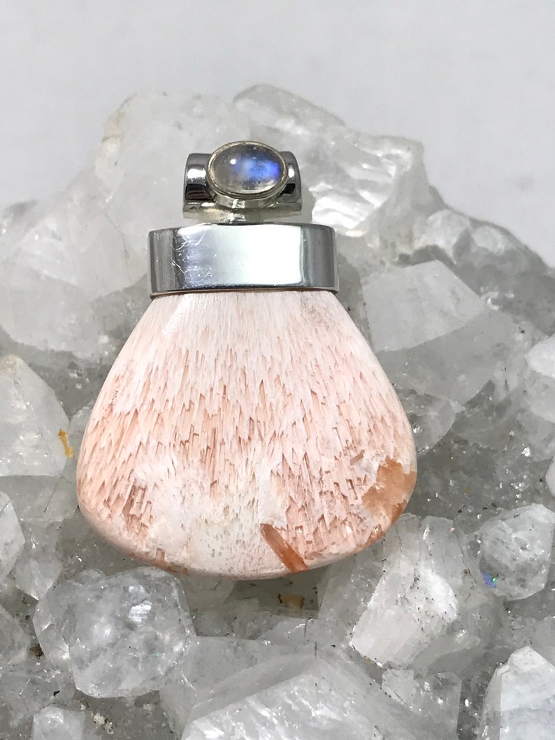 Op de afbeelding: Een zilveren hanger met een witte en roze edelsteen. De edelsteen is gezet in een zilveren bezel en heeft een moonstone cabochon bovenop. De hanger ligt op een bed van witte kristallen.