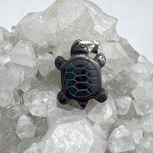 May include: A black turtle pendant charm made of polished obsidian. The pendant is sitting on a bed of white quartz crystals.