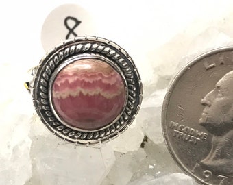 May include: A silver ring with a pink and white banded gemstone set in a rope-like border. The ring is next to a US quarter for size comparison.