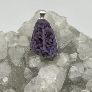 May include: A pendant featuring a polished, grape-like, purple druzy stone set in a silver-colored metal frame. The stone has a bubbly texture and a natural, organic shape. The pendant is displayed on a bed of clear quartz crystals.