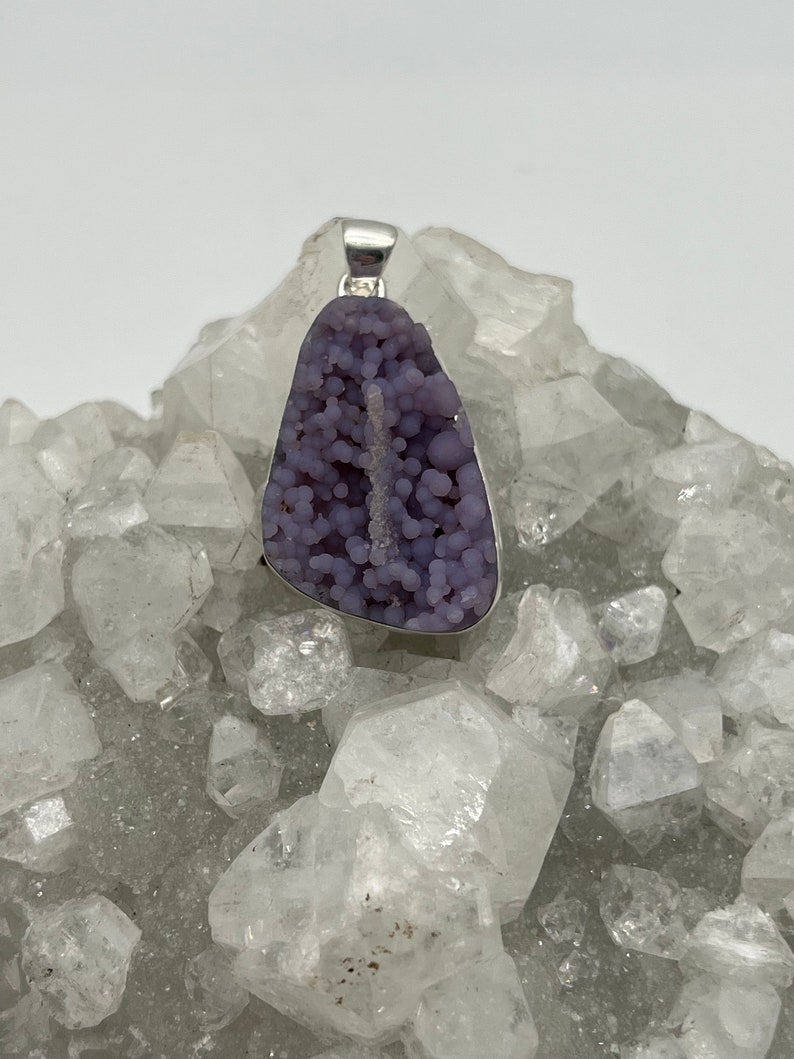 May include: A pendant featuring a polished, grape-like, purple druzy stone set in a silver-colored metal frame. The stone has a bubbly texture and a natural, organic shape. The pendant is displayed on a bed of clear quartz crystals.