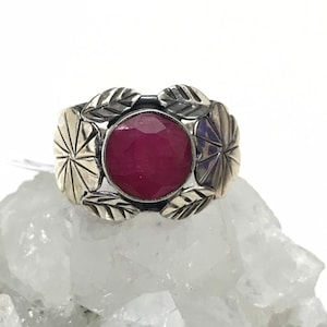 Puede incluir: Un anillo de plata con una gran gema de rubí redonda y facetada. El anillo está adornado con diseños de hojas y pétalos. El rubí es de color rojo intenso y el anillo está sobre un fondo blanco.