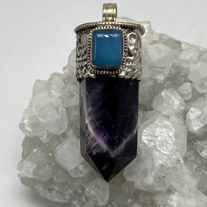 May include: A pendant featuring a polished amethyst crystal with a silver-coloured metal cap and bail. The cap has a square, light blue stone and decorative etched details. The crystal is a deep purple with white striations.