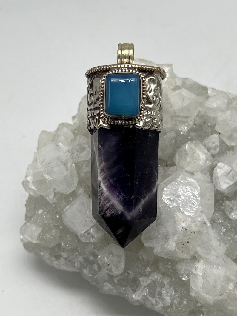 May include: A pendant featuring a polished amethyst crystal with a silver-coloured metal cap and bail. The cap has a square, light blue stone and decorative etched details. The crystal is a deep purple with white striations.