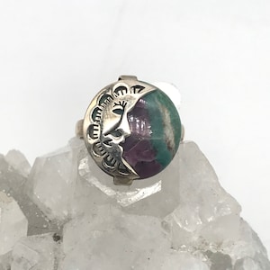 May include: A silver ring with a crescent moon design featuring a face and a green and purple gemstone.