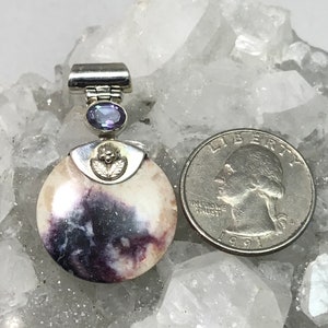 May include: A silver pendant featuring a round, white and purple stone with a silver bail and a small purple gemstone. The pendant is shown on a bed of white crystals.