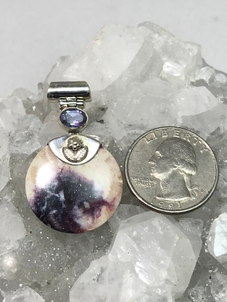 May include: A silver pendant featuring a round, white and purple stone with a silver bail and a small purple gemstone. The pendant is shown on a bed of white crystals.