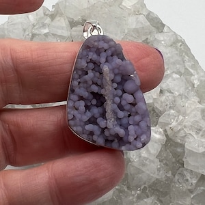 May include: A lavender-colored pendant with a silver bail. The pendant has a bubbly, textured surface. The gemstone is held by a silver bezel. The background is a white, crystalline surface.
