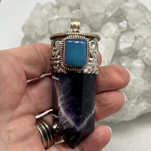 May include: A silver pendant with a rectangular blue gemstone and a carved silver design. The pendant is attached to a purple and black crystal. The pendant is held in a hand.
