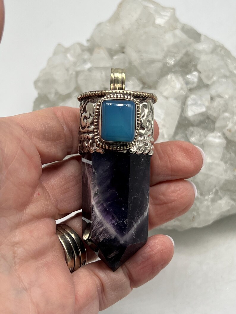 May include: A silver pendant with a rectangular blue gemstone and a carved silver design. The pendant is attached to a purple and black crystal. The pendant is held in a hand.