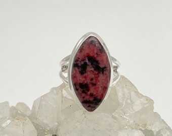 May include: A silver ring featuring a large, oval-shaped gemstone. The gemstone is a mix of deep red and black hues. The ring has a double-band design on the sides, adding a touch of elegance to the jewelry.